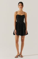 A woman is wearing a black dress with thin spaghetti straps. The dress has a fitted bodice with a slightly ruched design and a flared, above-the-knee skirt. She is also wearing black open-toe sandals with a small heel. Her accessories include gold earrings and rings. Her hair is styled in a sleek, low bun, and she has a neutral facial expression.